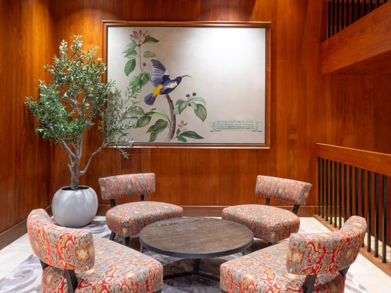 An intimate seating area in the lobby offers upholstered chairs around a circular table, framed by botanical artwork and wood walls, accented with blush patterns, olive greens, and warm walnut tones.
