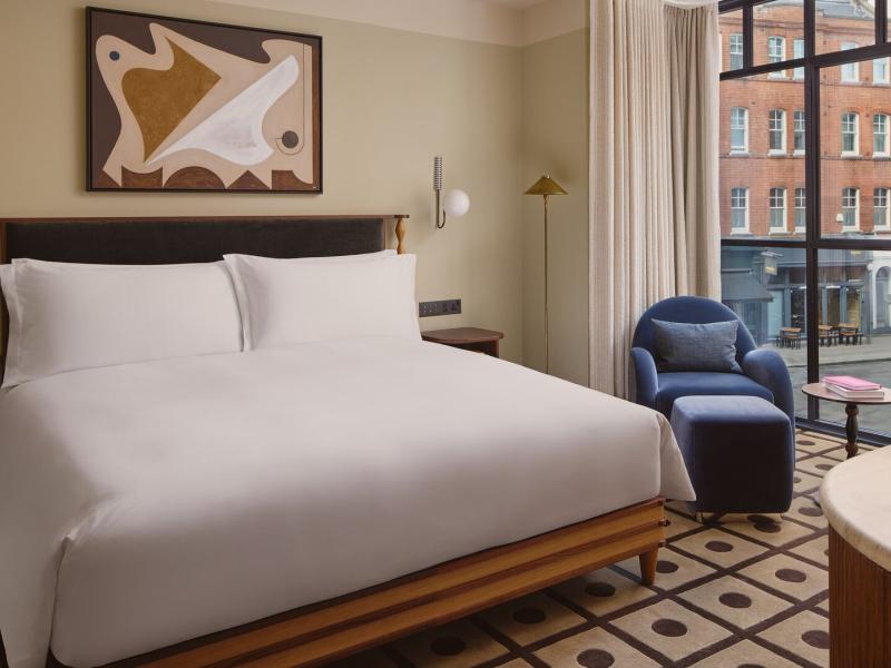 A bright One-Bedroom Suite features a plush king bed, large street-facing windows, and modern artwork, accented with warm wood tones, soft beige, and hints of navy blue.