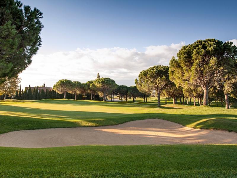 This scenic golf course fairway at Hotel Peralada showcases perfectly manicured emerald grass and a smooth sandy bunker, set against tall pine trees and a golden late‑afternoon sky.