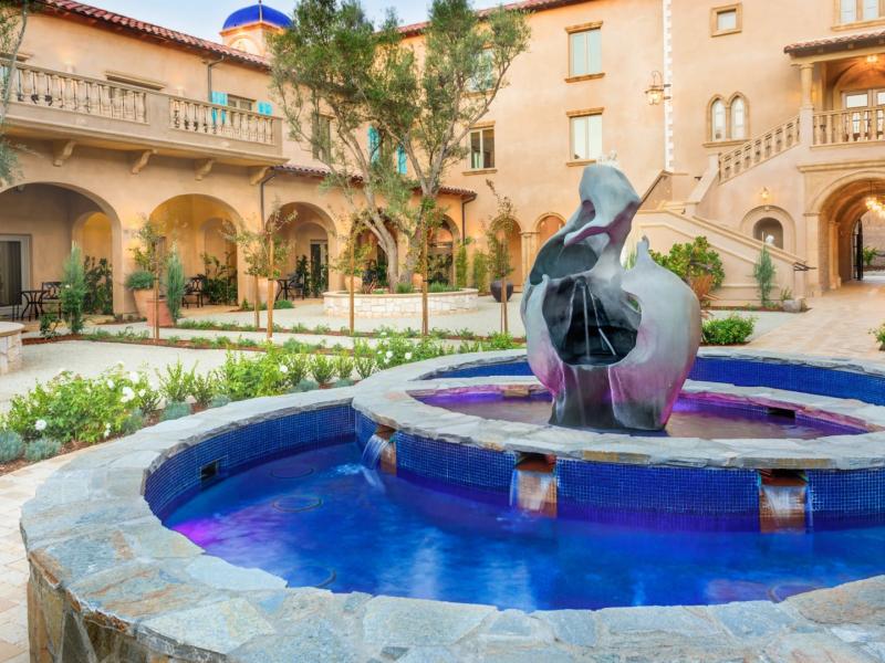 A sculptural centerpiece fountain anchors the Piazza Magica Courtyard with flowing forms and tiered stonework, brought to life by vibrant cobalt blue water, soft gray stone, and verdant greenery.