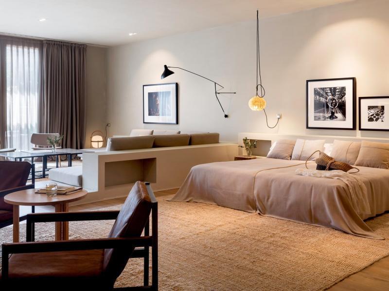Hotel Peralada spacious Presidential Suite features a soft beige palette with warm wood accents, highlighted by modern black lighting fixtures and floor‑to‑ceiling taupe drapes.
