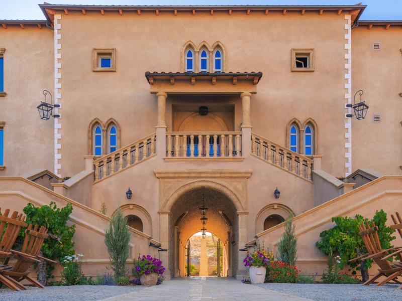 In the Piazza Magica Courtyard of Allegretto Vineyard Resort, an elegant, symmetrical façade features a dramatic central staircase, arched passageway, and wrought-iron details highlighted by sandy stone, muted blue windows, and natural wood accents.