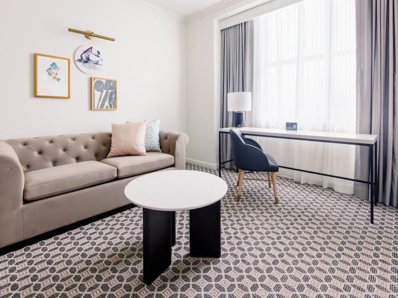 The Signature Suite bright sitting area showcases a tufted sofa and spacious work desk accented with soft blush tones and deep charcoal details.