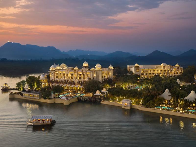 The Leela Palace Udaipur majestic lakeside palace glows at dusk with illuminated domes and terraces, accented by warm gold lighting, soft amber hues, and deep blue waters.
