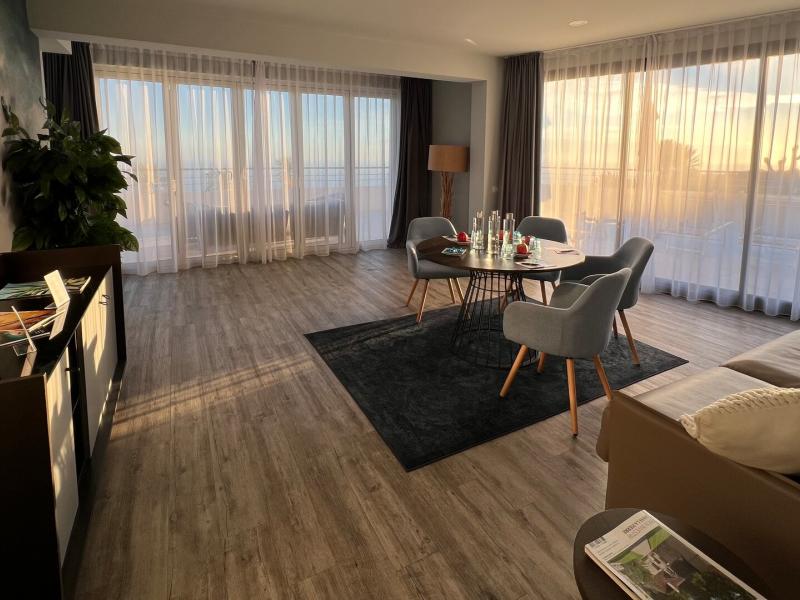 Hotel Les Terrasses D'Eze’s bright, modern meeting lounge featuring floor‑to‑ceiling windows, a cozy seating area, and a central table, accented with soft gray furnishings and warm wood tones.