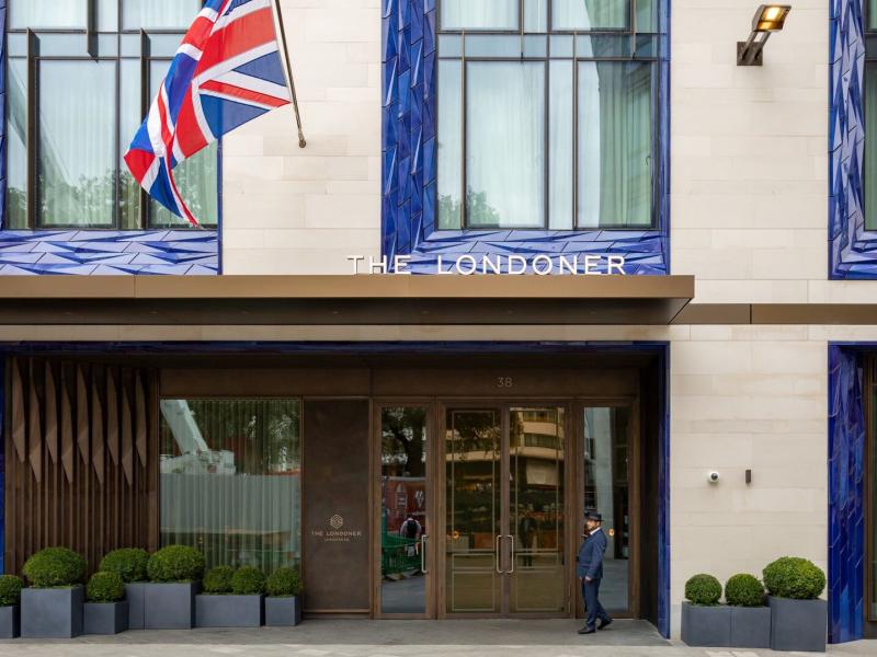 The Londoner’s stunning façade of cream stone and cobalt-blue tile frames pairs with a brass canopy, manicured greenery, and the iconic red, white, and blue Union Jack for a polished London arrival.