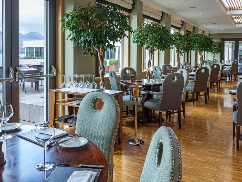 The Brasserie Restaurant elegantly pairs polished wood floors and serene lake views with refined table settings and sculpted greenery, accented in soft sage green, warm oak, and brushed silver tones.