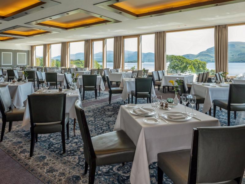 The Panorama Restaurant dining room showcases floor-to-ceiling windows, crisp white linens, and open lake and mountain vistas, enriched with cool slate gray, soft ivory, and warm amber lighting accents.