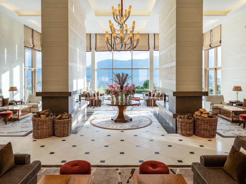 The Europe Hotel & Resort soaring, light-filled lobby showcases elegant neutral stone, sculptural décor, and plush seating accented with warm beige, soft taupe, and rich terracotta tones beneath a statement chandelier and panoramic mountain views.