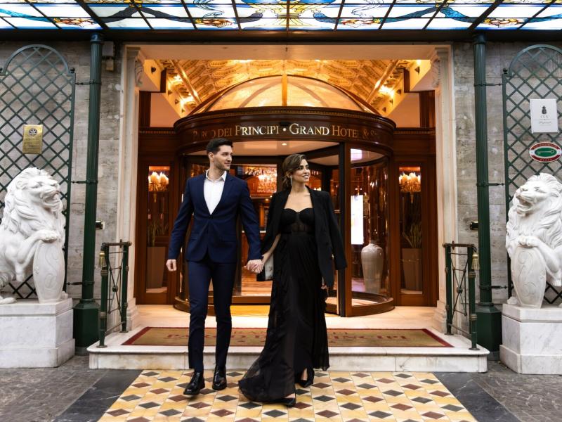 Parco dei Principi Grand Hotel & Spa’s elegant grand entrance showcases a gleaming revolving door, mosaic marble flooring, and classic stone lions, highlighted by warm gold lighting with black and cream accents.