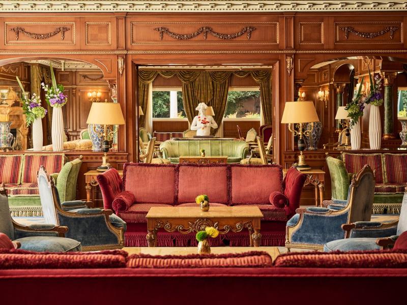The luxurious lobby lounge of Parco dei Principi Grand Hotel & Spa displays ornate wood paneling, mirrored walls, and plush seating, enriched with burgundy, emerald green, and gold accents for a timeless, opulent feel.