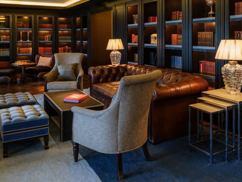 The intimate Library features dark wood bookcases, classic leather seating, and soft lamp lighting, layered in deep chocolate brown, charcoal, and warm honey-gold accents for timeless sophistication.
