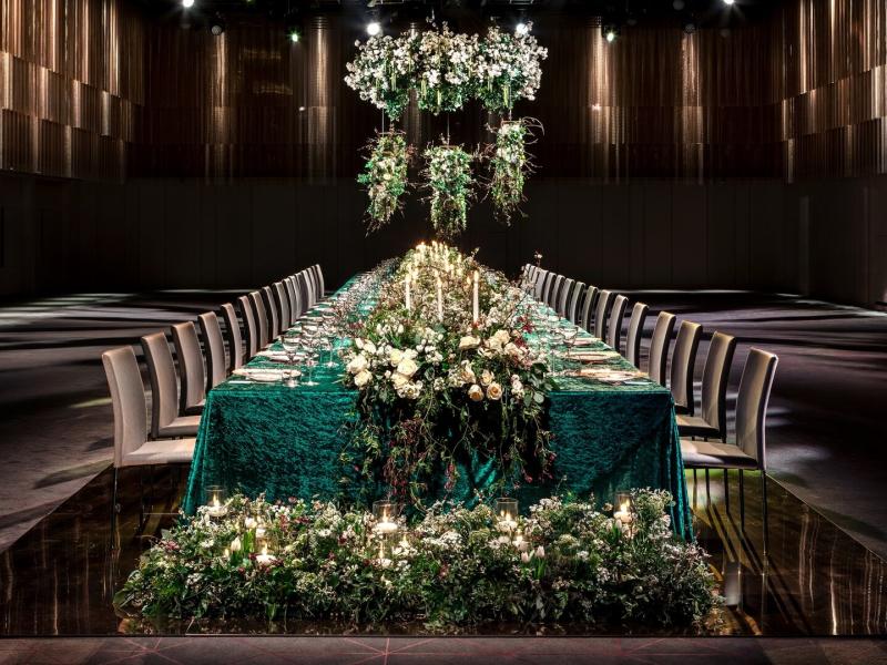 An elegant private dining table at the center of The Ballroom is dressed in emerald green linens, cascading florals, and candlelight, accented by deep green foliage and shimmering gold details.
