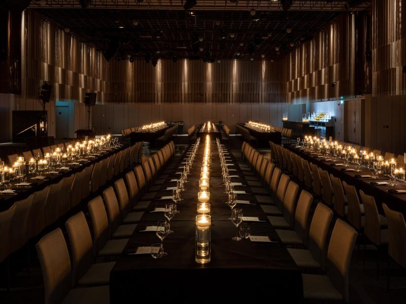 The Ballroom featuring a luxurious long-table dining experience glows with rows of candlelight, sleek black tabletops, and bronze-accented chairs in warm gold and soft mocha tones.