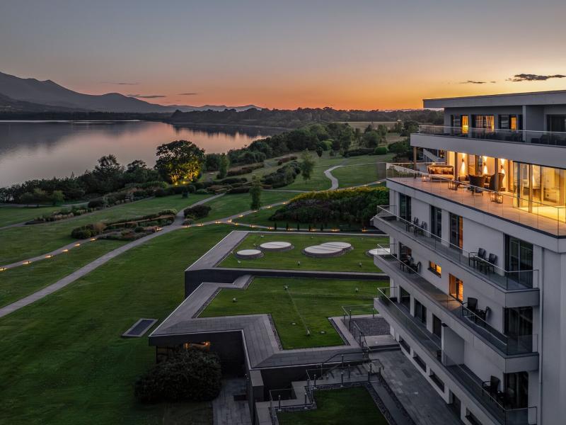 The Europe Hotel & Resort sleek lakeside retreat rises above manicured grounds and serene walking paths, glowing at sunset with warm amber lighting set against cool gray architecture and deep green landscapes.