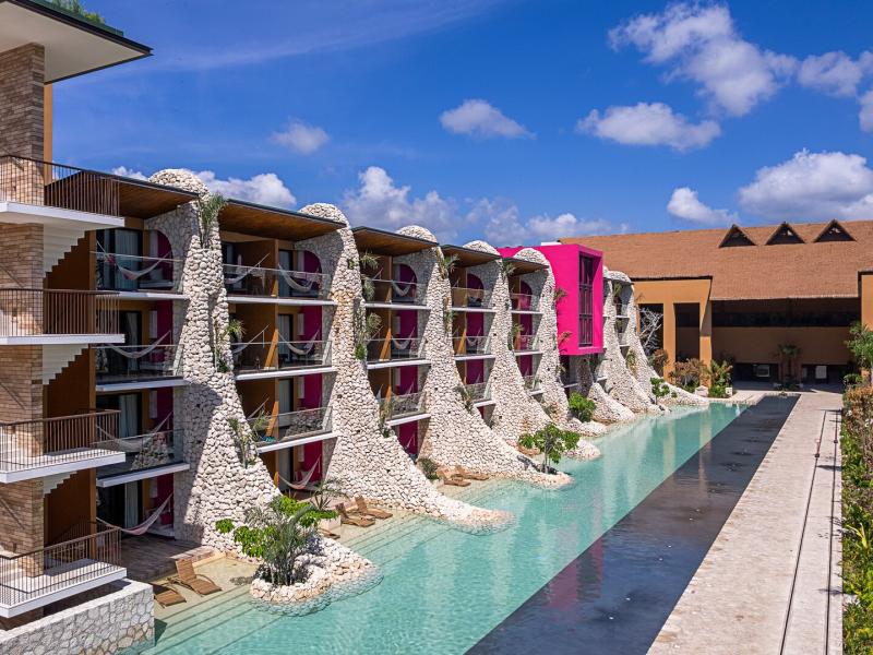 Striking stone-clad suites at Hotel Xcaret Mexico line a serene turquoise lagoon, featuring private balconies, sculptural facades, and bold accents of magenta, sandy beige, and aqua blue.