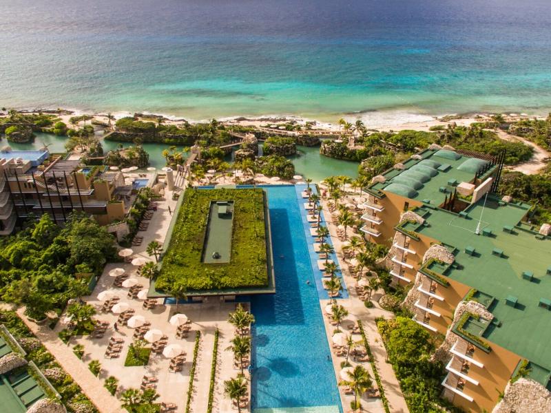 A stunning aerial view of Hotel Xcaret Mexico reveals infinity pools stretching toward the sea, surrounded by palm-lined decks and modern buildings in shades of blue, sandy beige, and lush green.