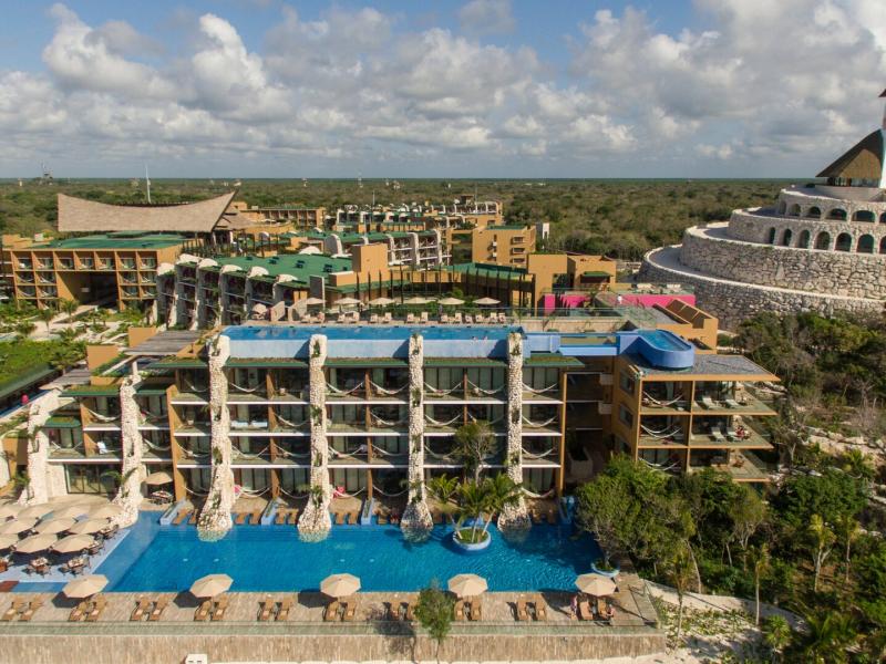 An elevated panoramic view of Hotel Xcaret Mexico showcases expansive resort buildings and a signature stone structure, accented by it’s Fuego and rooftop turquoise pools, and warm neutral architecture.