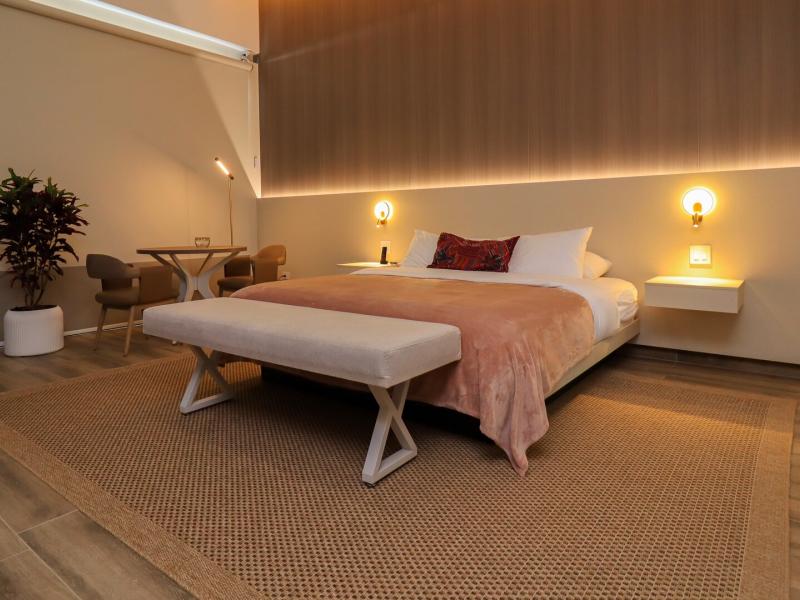 This spacious Canopy Suite bedroom showcases a minimalist platform king bed, integrated ambient lighting, and a cozy seating nook, finished in soothing taupe, warm wood tones, and muted rose accents.