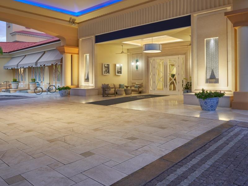 Eau Resort & Spa grand covered entrance showcases elegant columns, patterned stone pavers, and warm ambient lighting accented by soft sand tones, deep blue highlights, and lush green landscaping.