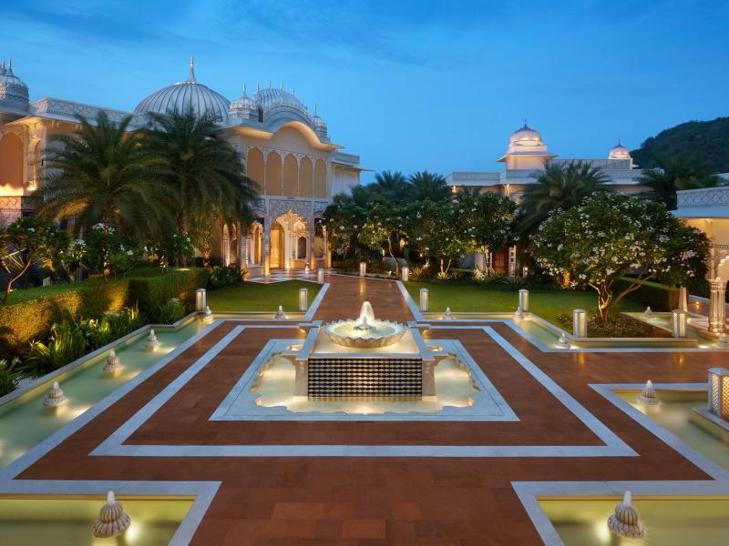 The grand courtyard of The Leela Palace Jaipur features a central illuminated fountain, symmetrical pathways, and it’s domed architecture highlighted by warm amber lighting, lush green landscaping, and terracotta accents.