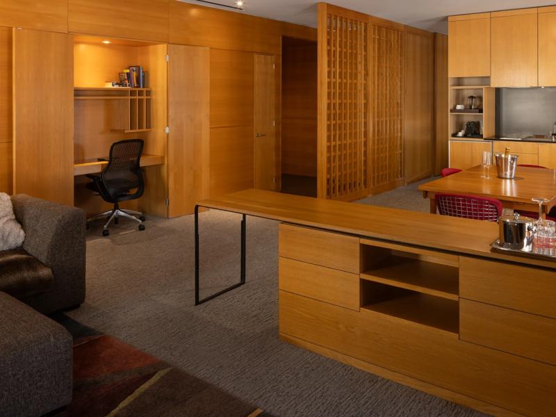 A spacious open‑concept Deluxe Apartment showcases custom wood cabinetry, a built‑in workspace, and a minimalist kitchen island, accented with honey-toned wood, charcoal gray upholstery, and subtle red details.