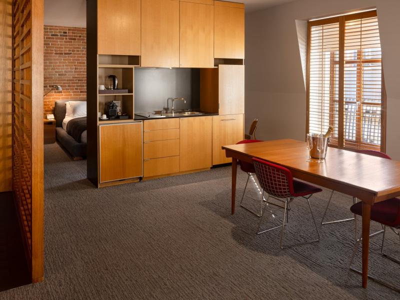 This dining and kitchen area fof a Deluxe Apartment at Hotel Gault highlights clean-lined cabinetry, a mid‑century dining table, and large shuttered windows, accented with light maple wood, muted gray carpeting, and pops of deep red seating.