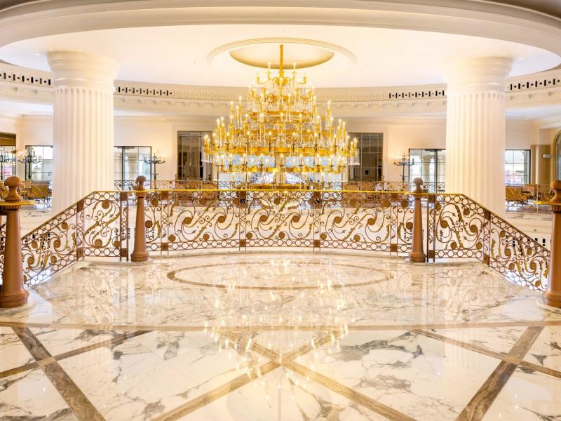 The lobby’s ornate gold ironwork frames a gleaming marble atrium, where a dramatic crystal chandelier glows against a refined palette of white stone, soft blush, and luminous gold.