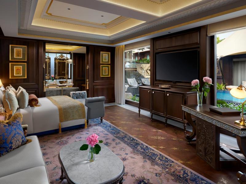 An elegant Leela Family Villa at Pichola bedroom features plush seating, a king bed, and poolside views, highlighted by rich dark wood, soft cream upholstery, and touches of royal blue and gold.