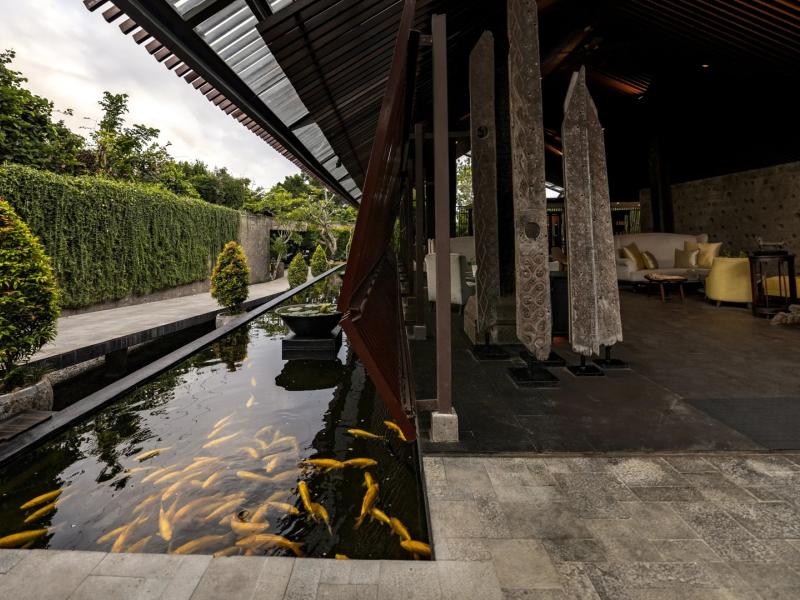 Ametis Villa Bali open-air lobby features a tranquil koi pond, sculptural stone pillars, and sleek teak surfaces accented by deep charcoal, warm beige, and flashes of golden orange.
