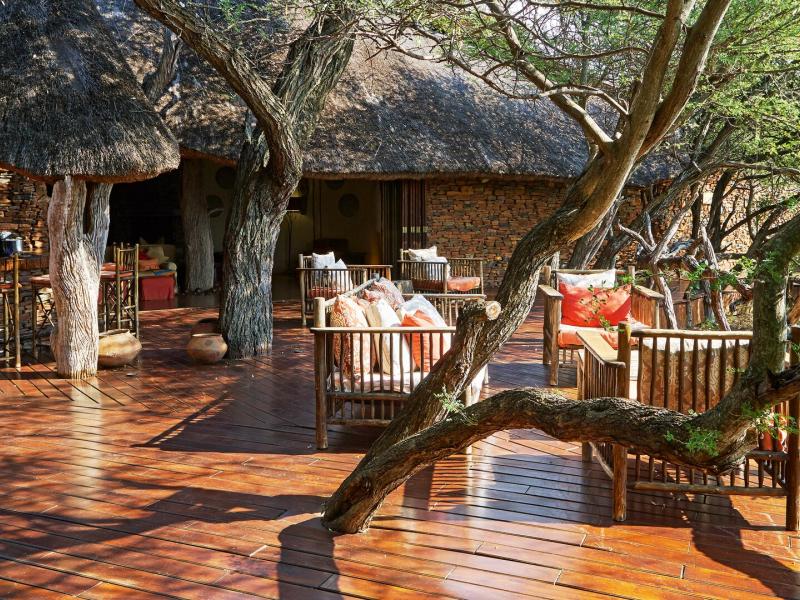 Last Word Makanyane expansive wooden deck set beneath a thatched roof features built‑in tree elements, rustic lounge seating, and warm accents of burnt orange and natural wood tones surrounded by lush green foliage.