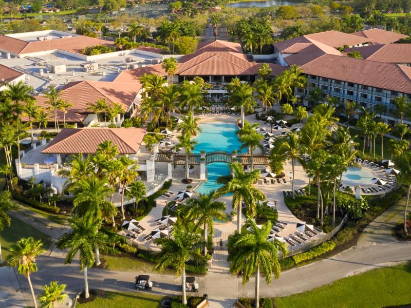 A sweeping aerial view reveals a palm-framed PGA National Resort with terracotta rooftops, serene turquoise pools, and manicured gardens woven through emerald landscapes.