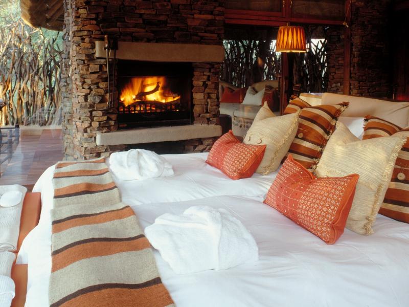 A luxurious open‑plan suite features a stone fireplace, plush white bedding, and layered textured pillows accented with warm rust, tan, and soft cream tones for a cozy yet refined retreat.