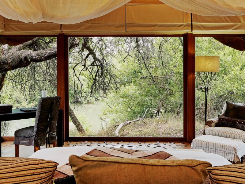 This airy suite features floor‑to‑ceiling windows, a cozy lounge area, and soft neutral furnishings accented with warm tan wood, muted cream, and earthy brown tones.
