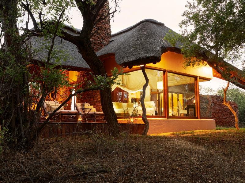 A suite exterior at Last Word Makanyane showcases a sweeping thatched roof, floor‑to‑ceiling glass windows, and a shaded terrace highlighted by earthy brown walls, soft amber lighting, and deep green surroundings.
