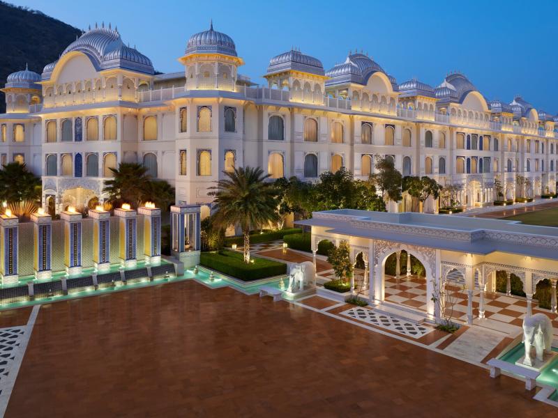 The majestic palace-style façade of The Leela Palace Jaipur displays ornate domes, arched windows, and cascading water features, glowing in soft ivory and gold with accents of teal and palm green.