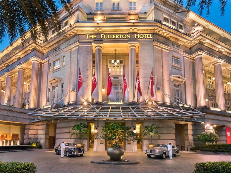 The Fullerton Hotel Singapore grand neoclassical façade with stately columns and a sweeping entrance is illuminated in warm golden light, accented by creamy stone details, rich red flags, and a deep blue evening sky.