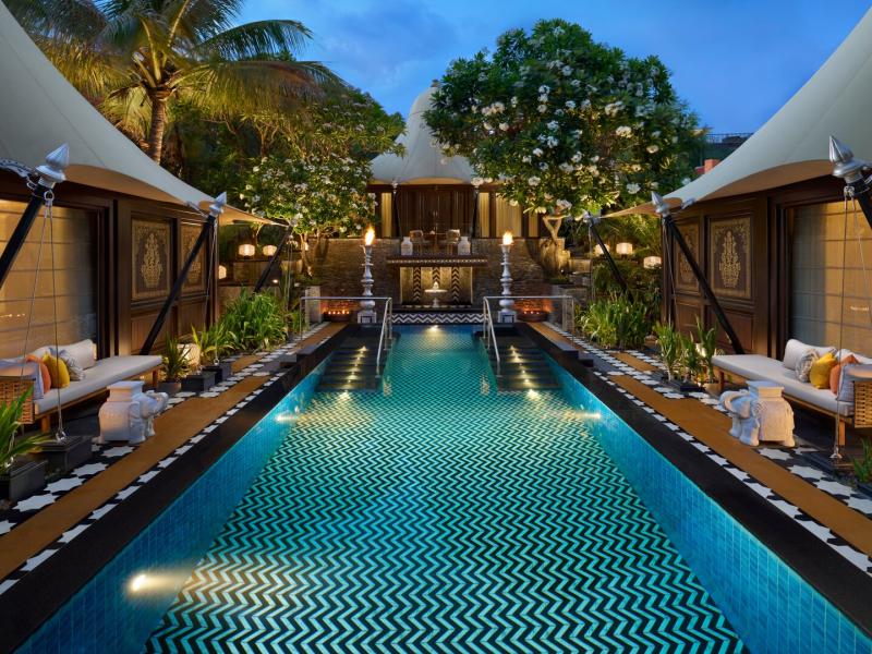 The Leela Family Villa’s serene private pool is framed by cabana-style seating, tropical landscaping, and patterned tiles, accented with teal blue water, lush green foliage, and warm golden lighting