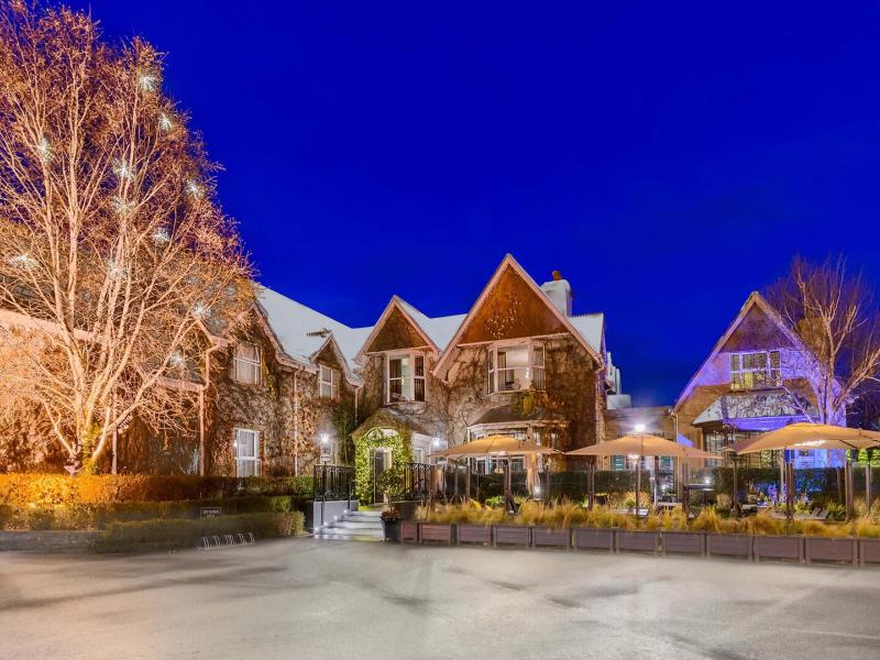 At twilight, The Victoria Killarney gabled exterior glows with warm ambient lighting and a chic terrace of canopy umbrellas, blending golden amber accents with rich stone, emerald foliage, and a deep sapphire sky.