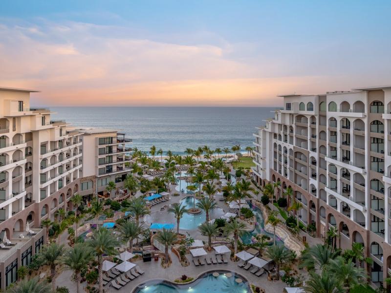 The striking Villa La Valencia Beach Resort & Spa Los Cabos oceanfront view showcases symmetrical modern architecture framing tiered infinity pools, lush palm-lined terraces, and cozy lounge seating accented in turquoise blue and warm sandy beige against the deep blue sea.
