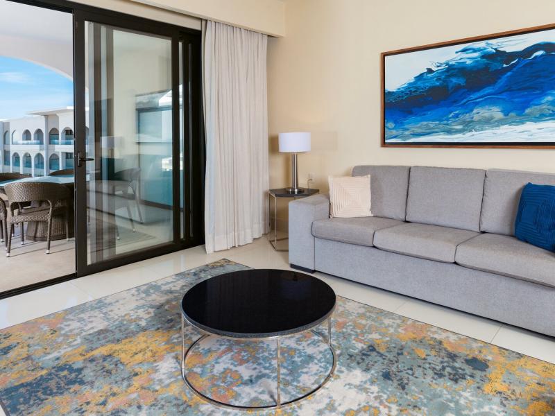 A Two Bedroom Suite Ocean View living area features a plush gray sofa, sliding glass doors to an ocean-view balcony, and layered décor accented in ocean blue, soft beige, and warm wood tones.