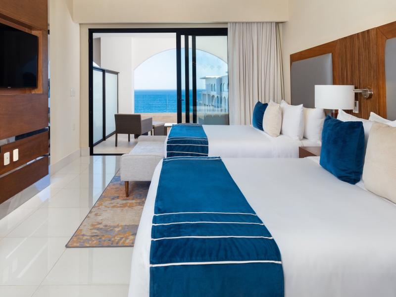This airy Two Bedroom Suite Ocean View at Villa La Valencia Beach Resort & Spa highlights floor‑to‑ceiling glass doors, a private balcony view, and tailored bedding with bold navy blue, ivory, and natural wood accents.
