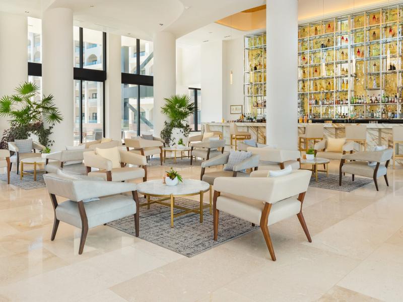 Villa La Valencia Beach Resort & Spa elegant lobby bar, La Jolla, features soaring windows, plush lounge seating, and a statement glass-backed bar, highlighted by soft ivory, warm gold, and fresh green accents for a bright, upscale ambiance.