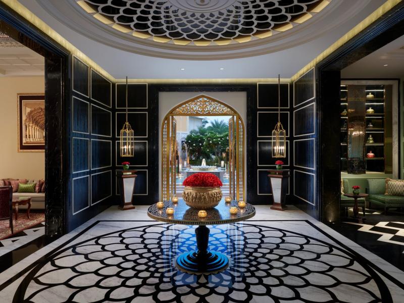 The Leela Palace Jaipur dramatic interior corridor impresses with a circular patterned floor, arched doorway, and sculptural centerpiece, contrasted by sleek black panels, warm gold lighting, and bold red accents.