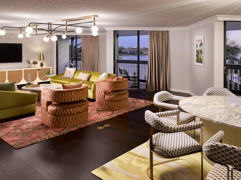 A sophisticated living and dining space of the Presidential Suite features plush olive and blush seating, a sculptural marble dining table, and warm brass lighting accents framed by expansive waterfront views at dusk.