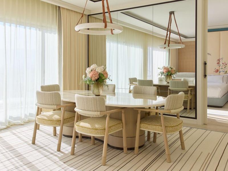 A Presidential Penthouse Suite light‑filled dining space showcases a sculptural white table, upholstered chairs, and mirrored walls in layered tones of ivory, sand, and soft champagne.