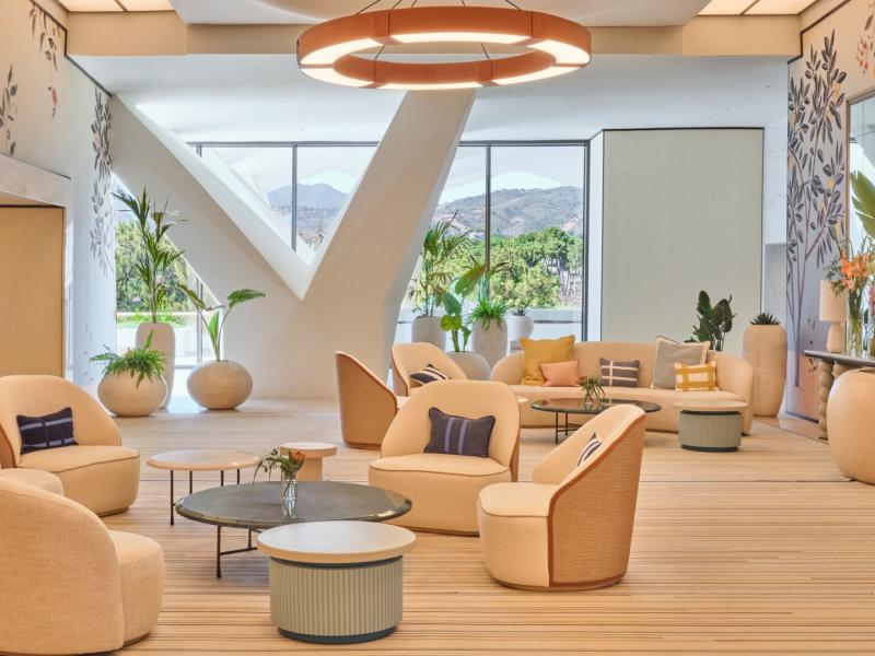 Don Carlos Marbella airy lobby lounge features curved upholstered seating, natural wood textures, and floor-to-ceiling windows accented with warm neutrals, soft gold, and botanical greens.