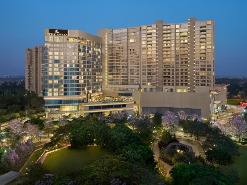 The Leela Bhartiya City striking high-rise exterior features sleek modern architecture and expansive glass façades, illuminated by soft golden lighting against cool blue skies and lush green surroundings.