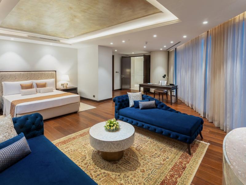The opulent Presidential Suite pairs a statement bed with an elegant lounge area, distinguished by rich sapphire blue upholstery, warm bronze detailing, and lustrous wood finishes.
