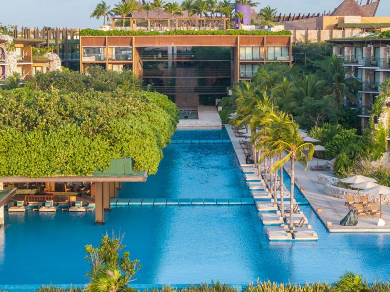 A luminous turquoise infinity pool bordered by swaying palms, plush ivory loungers, and a lush emerald-roofed bar is framed by rich honey and Hotel Xcaret Arte’s sand-hued architecture.